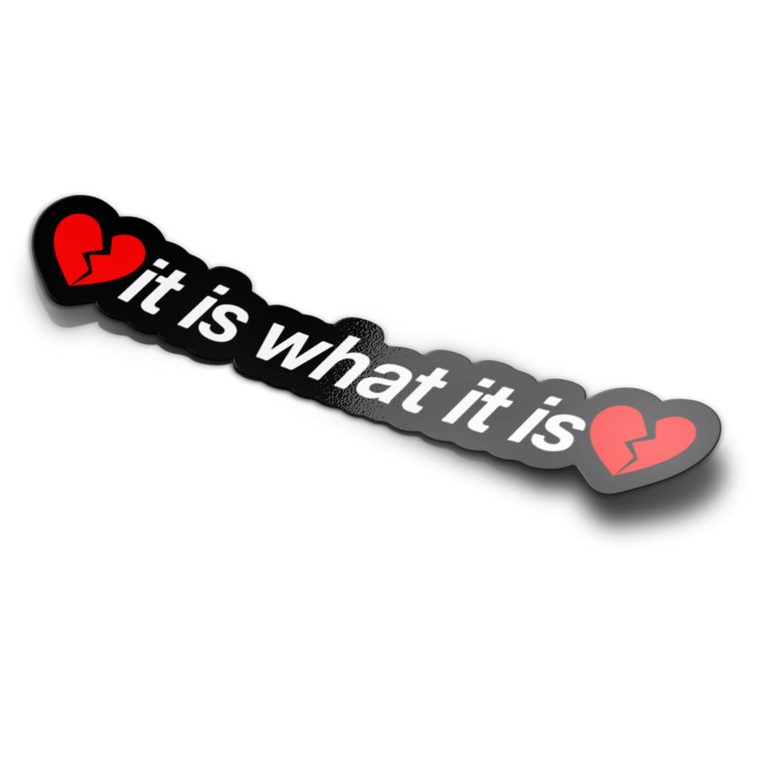 A sticker with a phrase 'it is what it is' written in a playful font, with a broken heart symbol both sides. The sticker is black with white text and red accents.